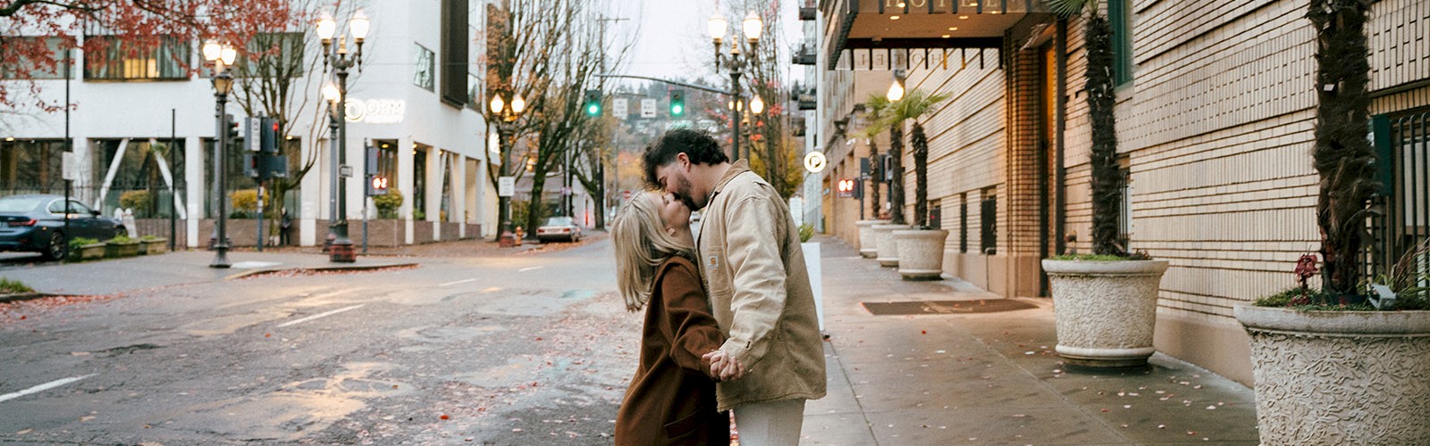 A couple shares a kiss on a city sidewalk near a hotel in a quiet urban street. The scene is calm with trees and an American flag visible.