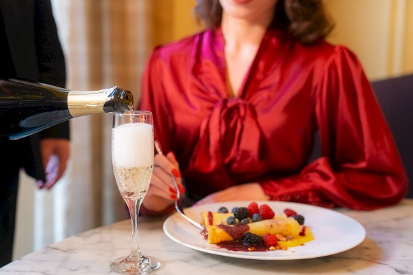 A person in a red blouse is having dessert with berries while a glass of champagne is being poured next to them on a marble table.