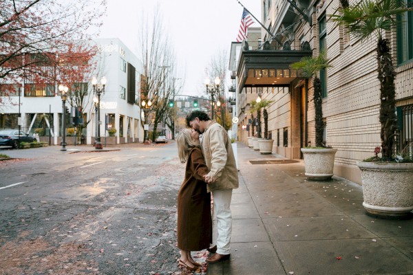 A couple is kissing on a city street near a hotel. The surroundings are lined with trees and buildings, with an American flag visible.