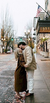 A couple stands on a city street, kissing in front of a building with the American flag, surrounded by trees and parked cars.