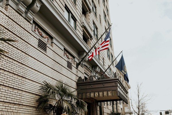 The image shows the exterior of a hotel building with two flags: the American flag and a state or organizational flag, against a cloudy sky.