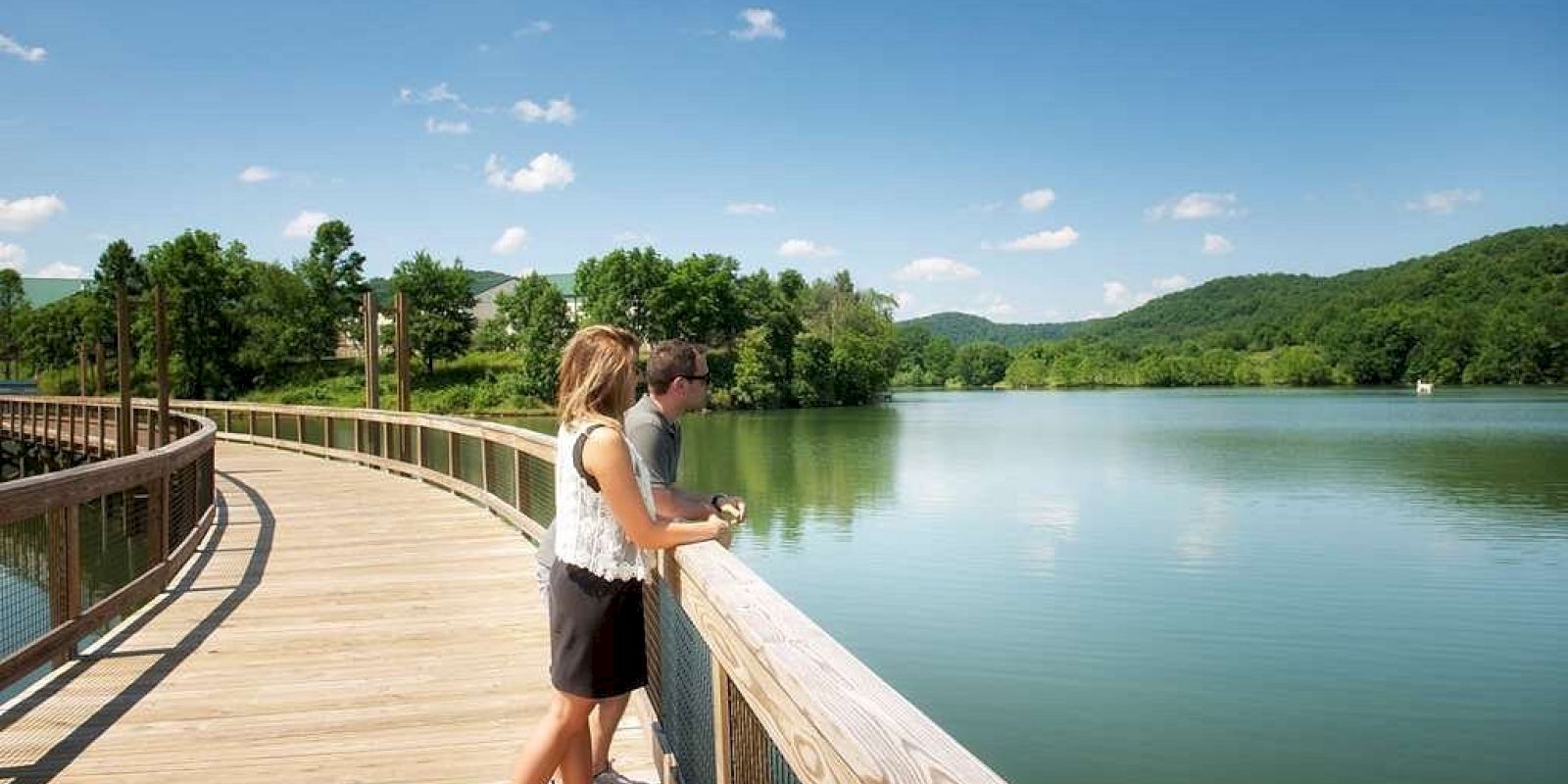 Two people stand on a wooden walkway by a serene lake, mountains in the distance, under a clear blue sky.