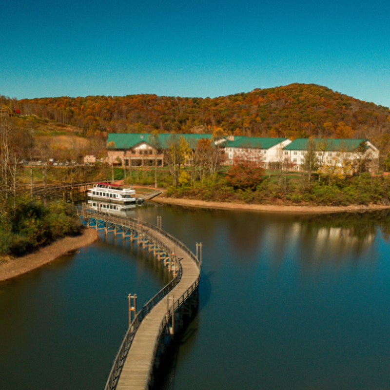 An aerial view of a serene lake with a curved boardwalk leading to a large building, surrounded by lush trees and hills under a clear blue sky.