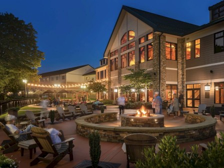 An evening scene of a large building with outdoor seating around a fire pit. String lights illuminate a patio area where people are gathered.