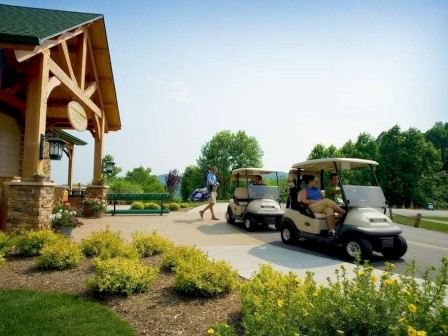 People are at a golf course with golf carts, one person walking with a golf bag, and a wooden building with greenery in the background.