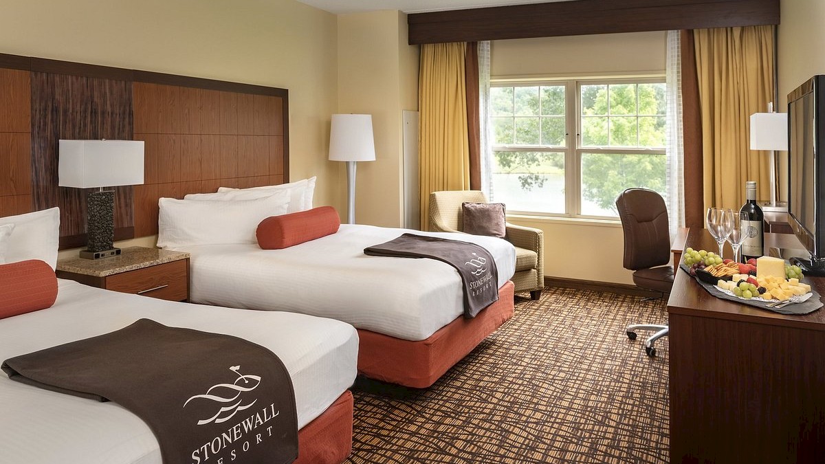 This image shows a hotel room with two beds, a desk with a chair, a window, and a table with wine and fruit.