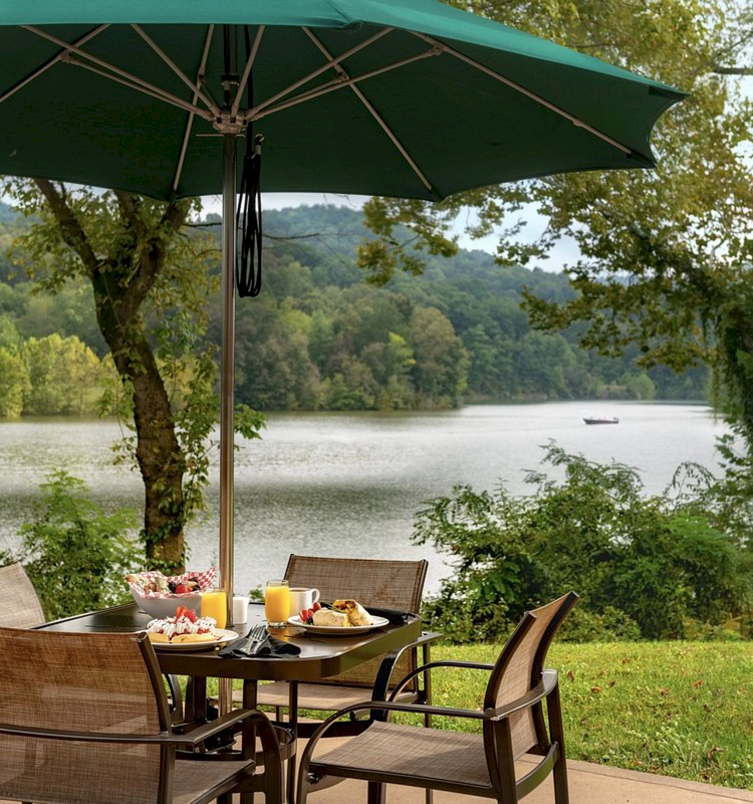 A table with chairs is set with food and drinks under a green umbrella, situated by a scenic lake and lush greenery in the background.