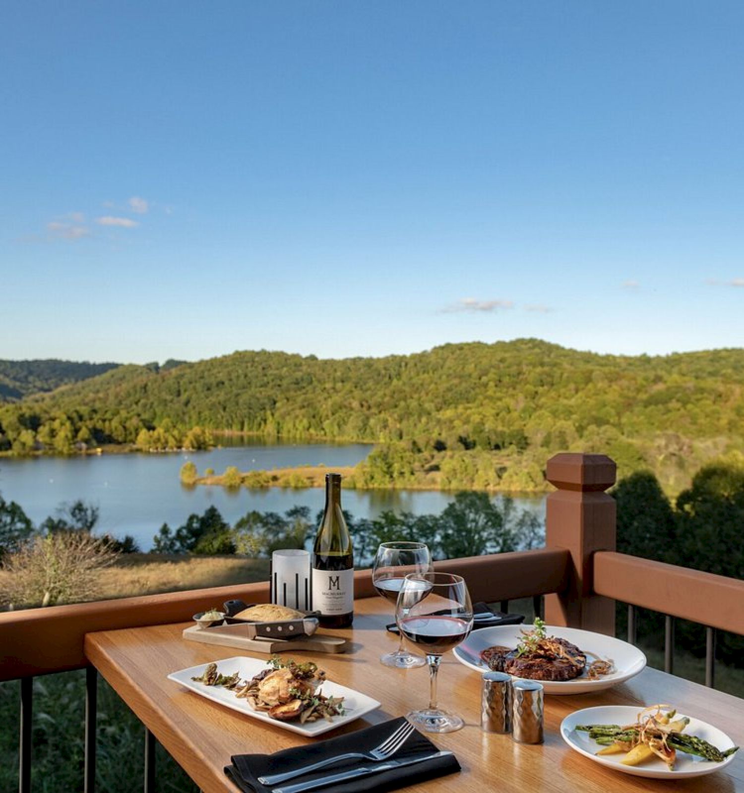 A table set for a meal overlooks a scenic lake and lush green hills under a clear blue sky, featuring wine and various dishes.