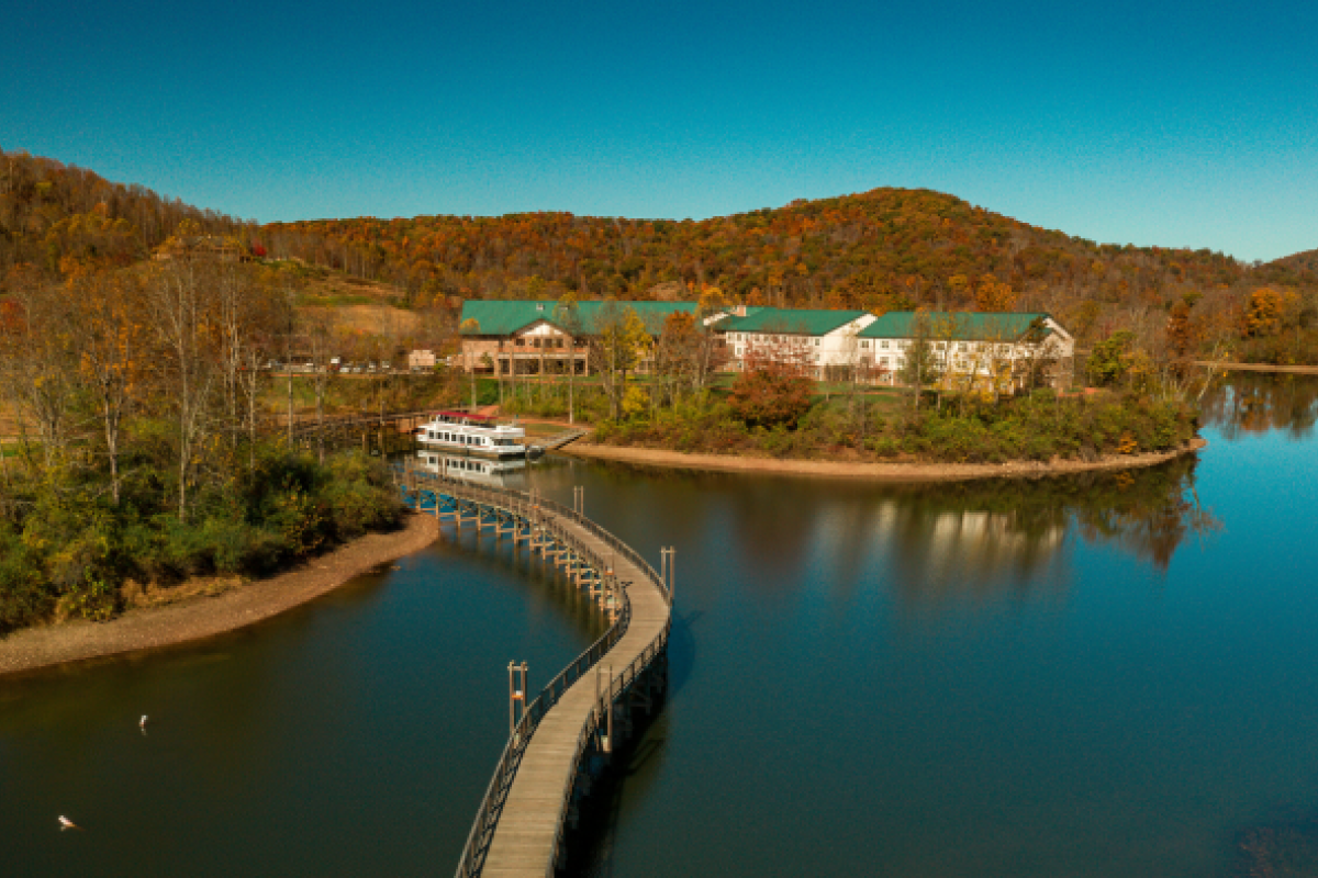 A scenic view of a lake with a long, winding boardwalk leading to a boat docked near buildings, surrounded by autumn-colored trees and hills.