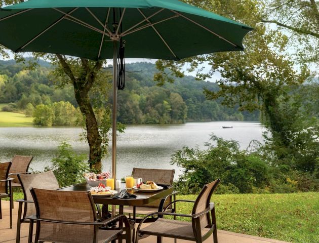 An outdoor dining setup with a table, chairs, and food under a green umbrella, overlooking a scenic lake and tree-covered hills in the background.