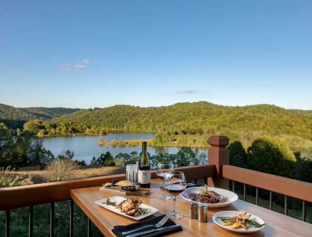 A table set for a meal with dishes and wine overlooks a beautiful lake and green hills under a clear blue sky.