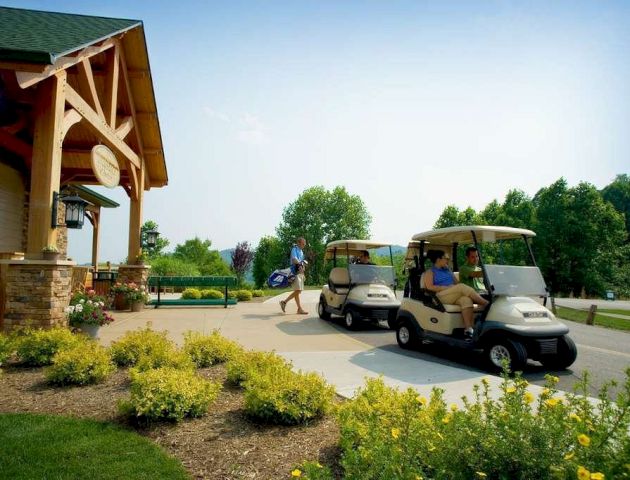 A person stands by a building while two others drive golf carts on a paved path surrounded by greenery and yellow flowers.