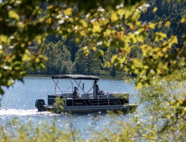 A pontoon boat with two people on board is cruising on a lake, surrounded by trees and greenery, on a sunny day.