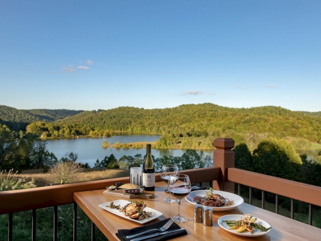 A picturesque outdoor dining setup featuring plates of food, wine, and glasses with a beautiful view of a lake and green hills in the background.