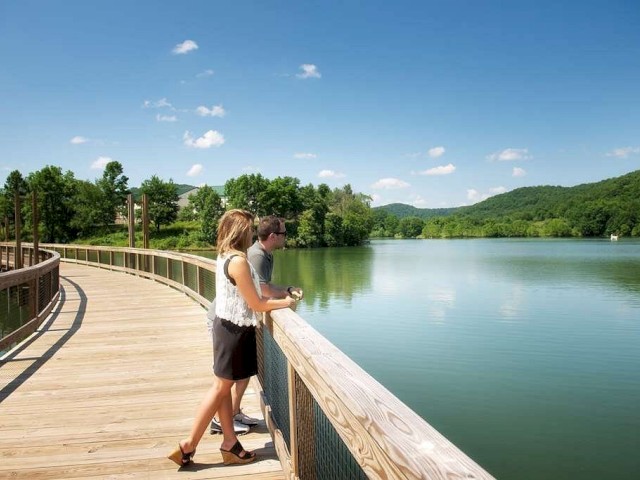 Two people standing on a wooden boardwalk overlooking a lake surrounded by greenery, under a clear blue sky.