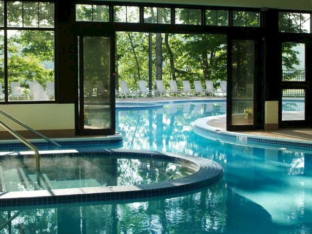An indoor pool area with large windows showing an outdoor pool and lounge chairs. The setting includes stairs and a separate hot tub section.