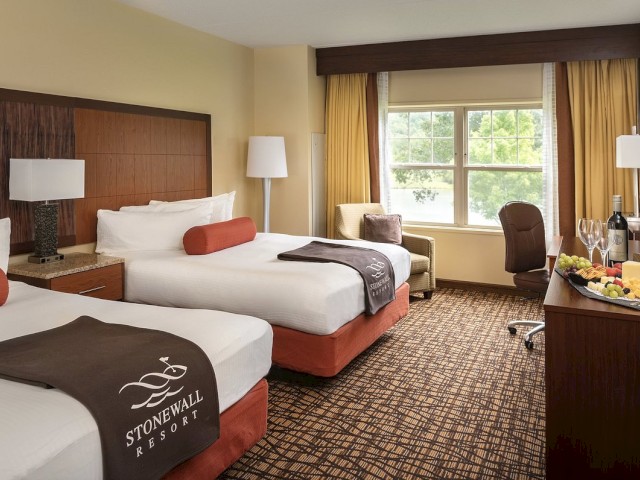 The image shows a hotel room with two beds, a window with yellow curtains, a small seating area, and a desk with wine and a fruit platter.
