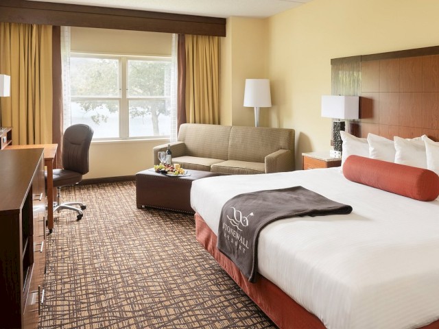 A tidy hotel room features a large bed, a sofa, a desk with a chair, a TV, and a coffee table, with natural light from a window.