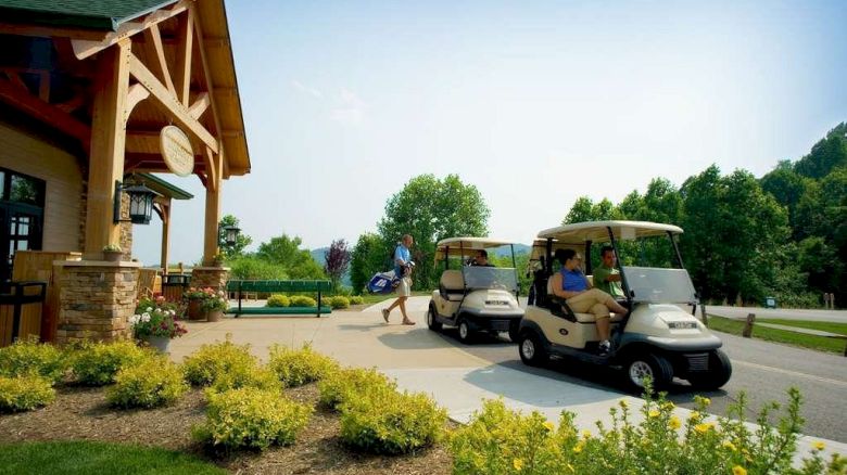 The image shows a person walking with a golf bag near a golf clubhouse, while two people sit in golf carts nearby, amid lush greenery and landscaping.