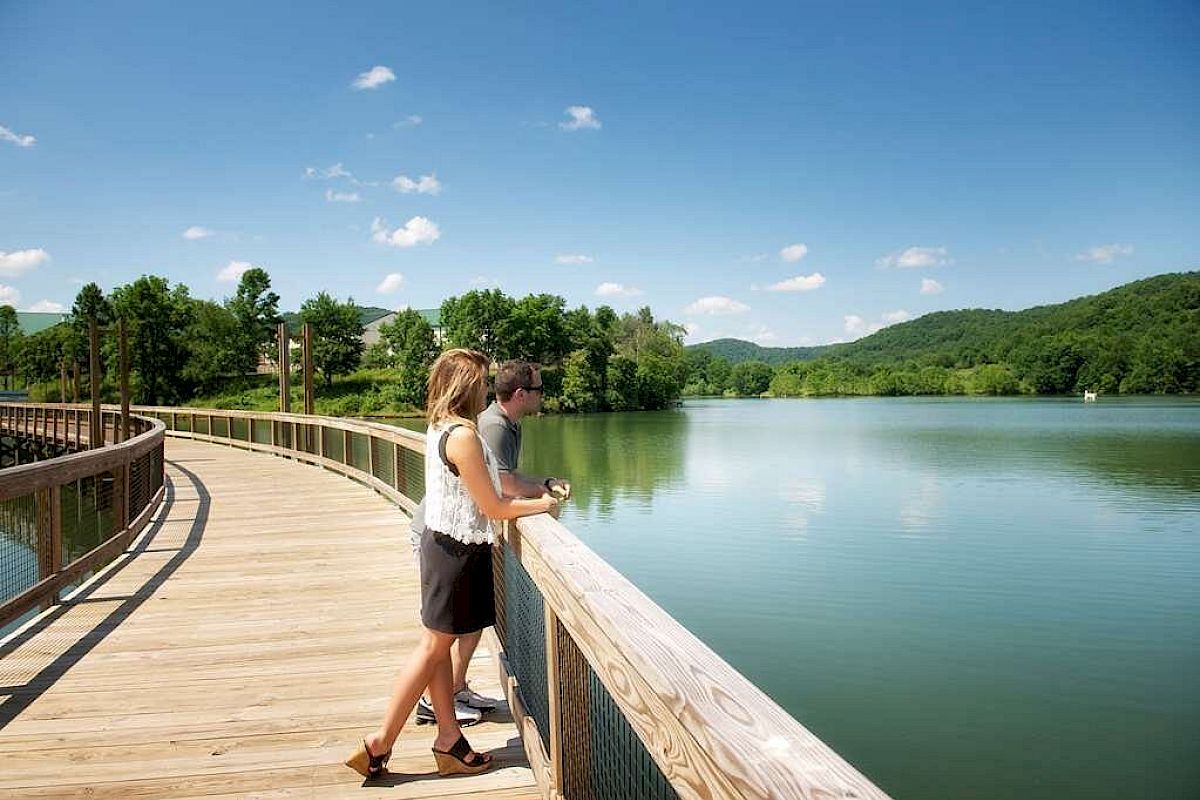 Two people stand on a wooden boardwalk overlooking a serene lake with lush green hills and trees in the background under a clear blue sky.