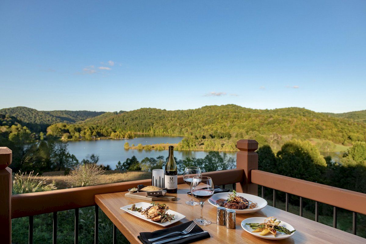 A table set with plates of food, wine, and glasses on a patio, overlooking a scenic lake and forested hills under a clear blue sky.